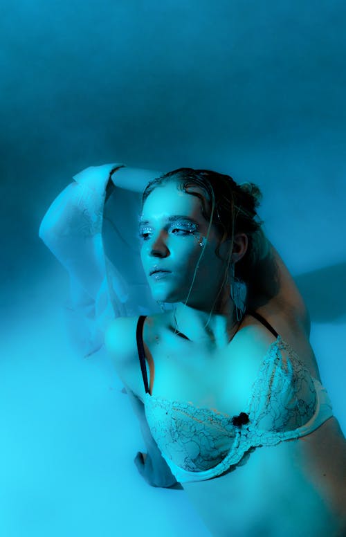 High angle seductive female in white bra sitting 9n floor in dark room in blue lights and looking away sensually
