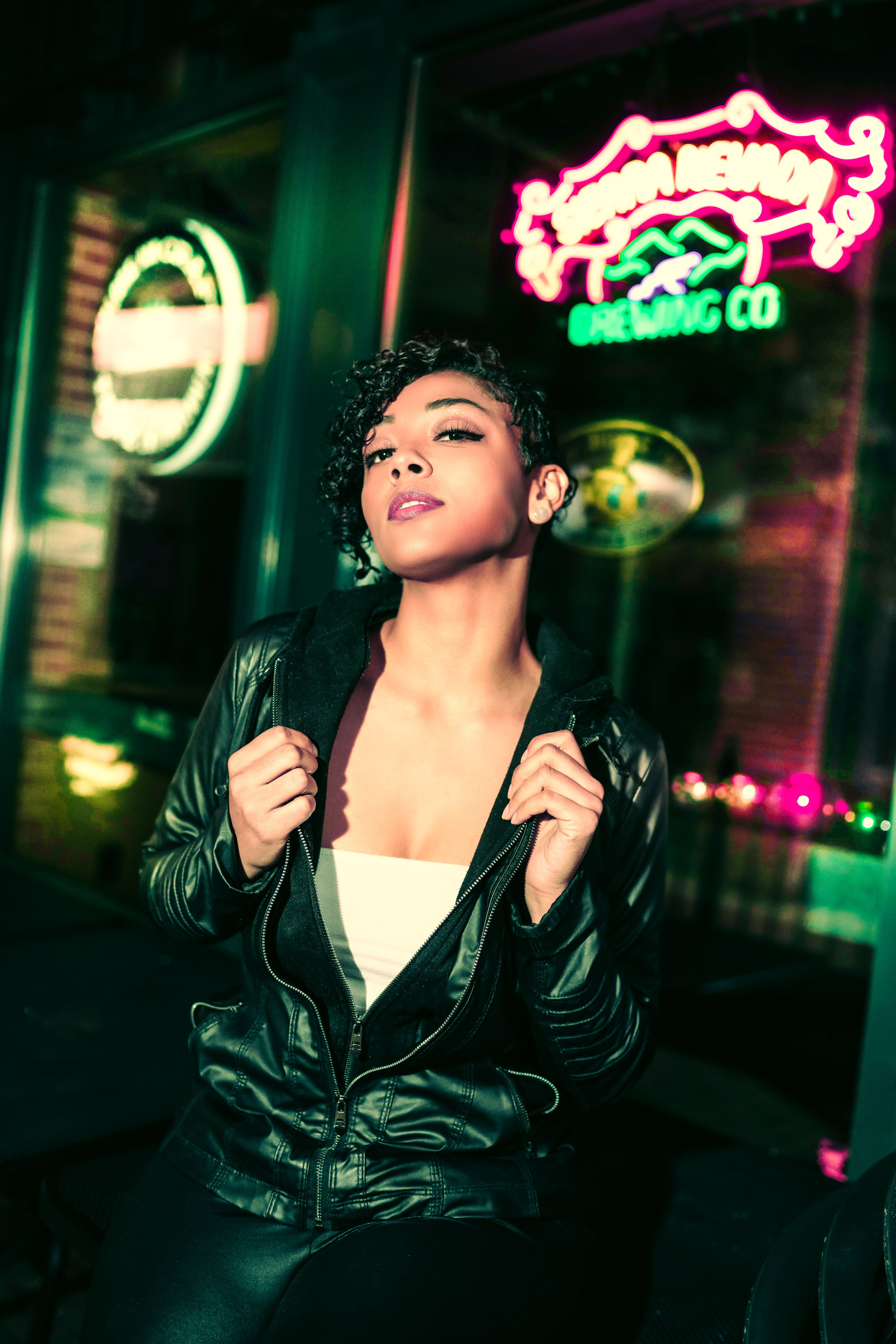 Confident young ethnic female wearing leather jacket and white top looking at camera while spending time near glowing neon bar sign in USA