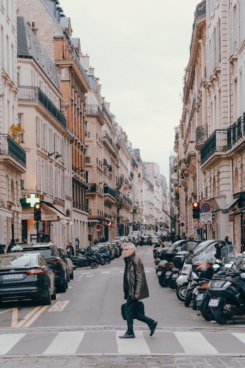 Full body side view of aged man in leather jacket crossing asphalt road with parked vehicles near buildings in city