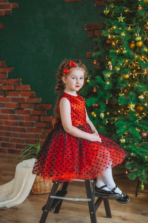 Full body of cute child in red dress relaxing on chair with crossed legs against Christmas tree and looking at camera