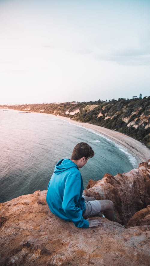 Free Photograph of a Man in a Blue Hoodie Sitting on a Cliff Near the Beach Stock Photo