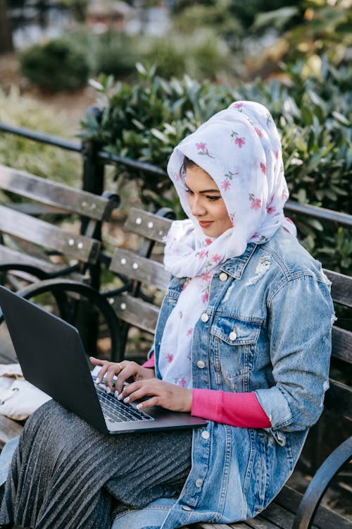 Free Smiling young Muslim female freelancer wearing trendy jeans jacket typing on netbook while sitting on shabby bench in park on summer day Stock Photo
