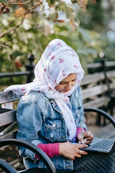Young Muslim woman in a hijab working remotely on her laptop outdoors in a park setting.