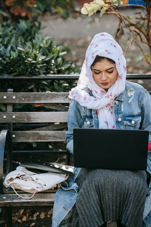Free Focused Muslim lady wearing skirt and floral headscarf sitting on old wooden bench in garden and using netbook for freelance work on sunny day Stock Photo