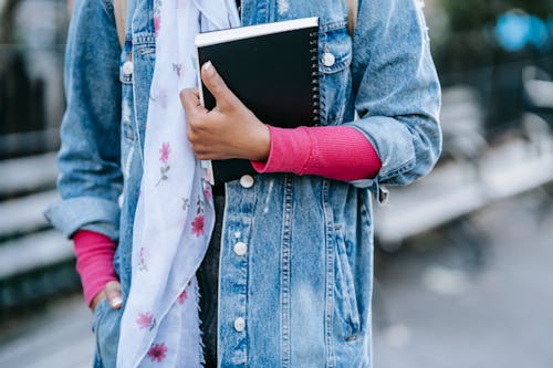 Crop anonymous woman wearing denim jacket and pink pullover with scarf and backpack holding black copybook while standing in park