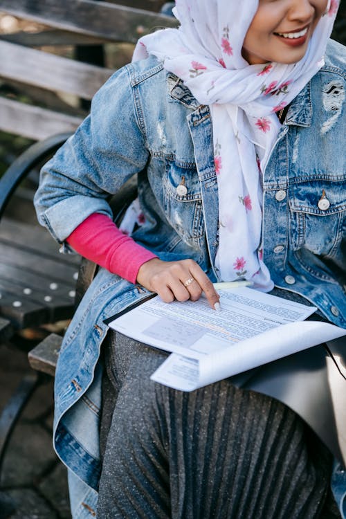 From above of crop smiling Muslim female entrepreneur checking information in project documents while sitting on wooden bench on sunny day