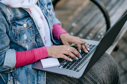 High angle of crop anonymous female freelancer wearing scarf sitting on wooden bench in park and holding pen in left hand and typing on laptop