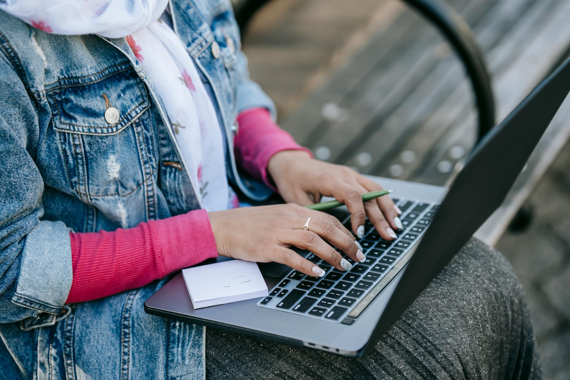 High angle of crop anonymous female freelancer wearing scarf sitting on wooden bench in park and holding pen in left hand and typing on laptop