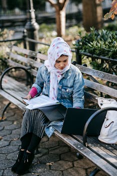 Concentrated young Muslim businesswoman wearing floral headscarf and denim coat with long skirt sitting on wooden bench in park and browsing laptop