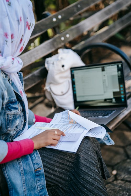 Crop woman in headscarf sitting on bench with paperwork and laptop