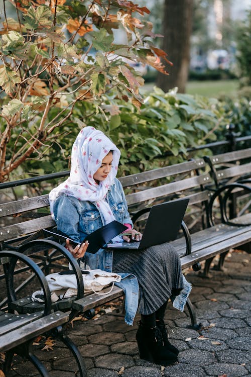 Concentrated young Muslim lady wearing headscarf and jeans coat with skirt surfing laptop while sitting with working documents on bench in park on sunny day