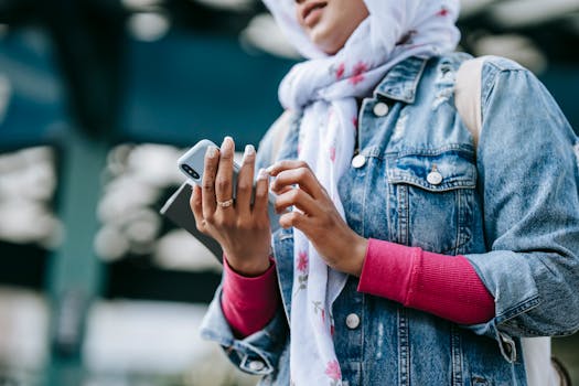 A young Muslim woman in a denim jacket and hijab texting on her smartphone in an urban outdoor setting.