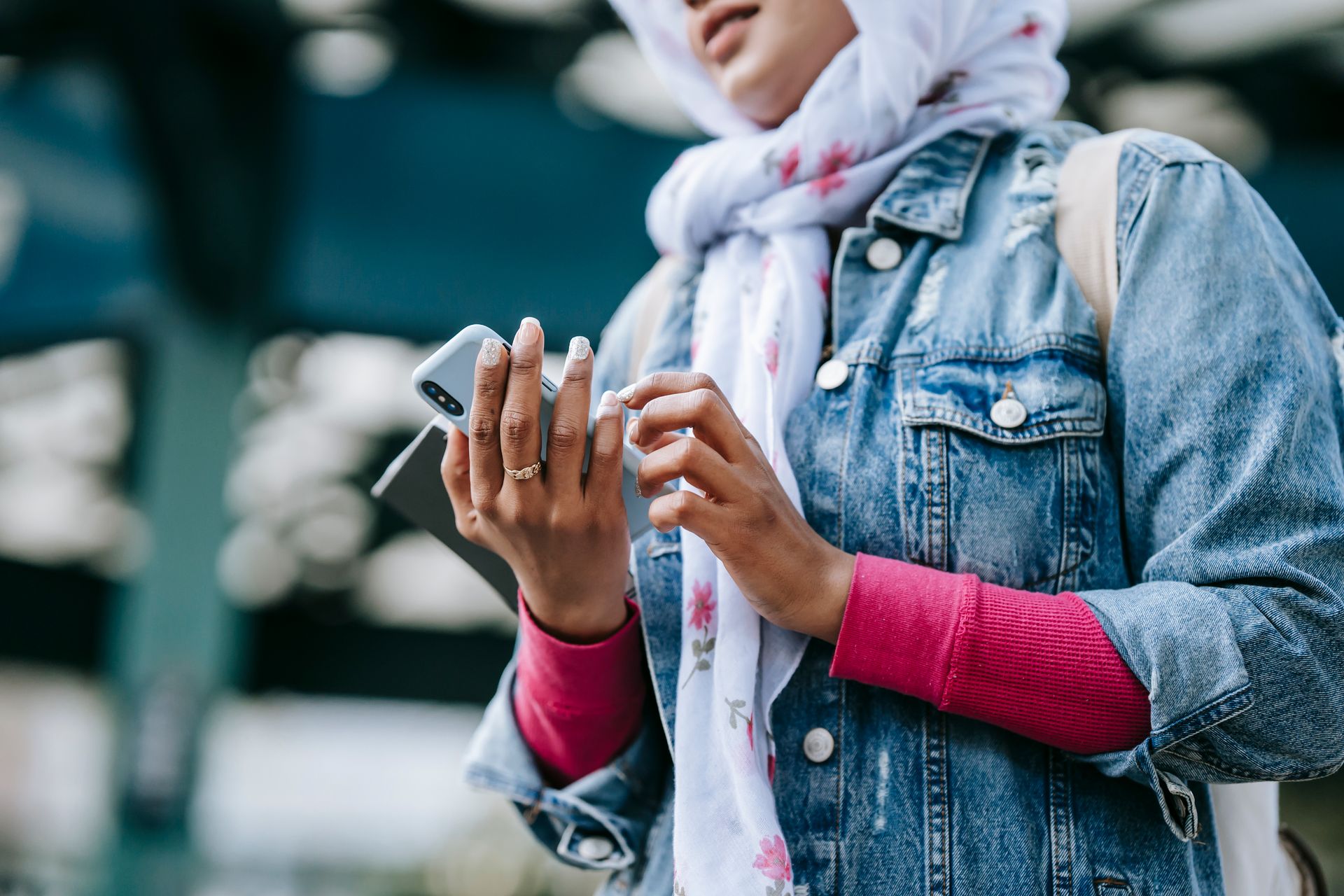 A young Muslim woman in a denim jacket and hijab texting on her smartphone in an urban outdoor setting.