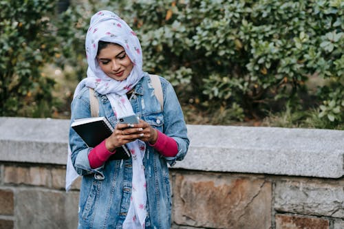 Cheerful young Muslim lady in hijab and jeans coat standing near stone fence of city park and surfing modern cellphone