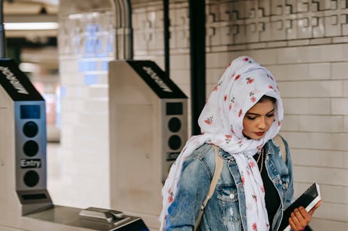 Calm Muslim woman with copybook wearing floral headscarf wearing jeans jacket with backpack entering subway station through automatic turnstile