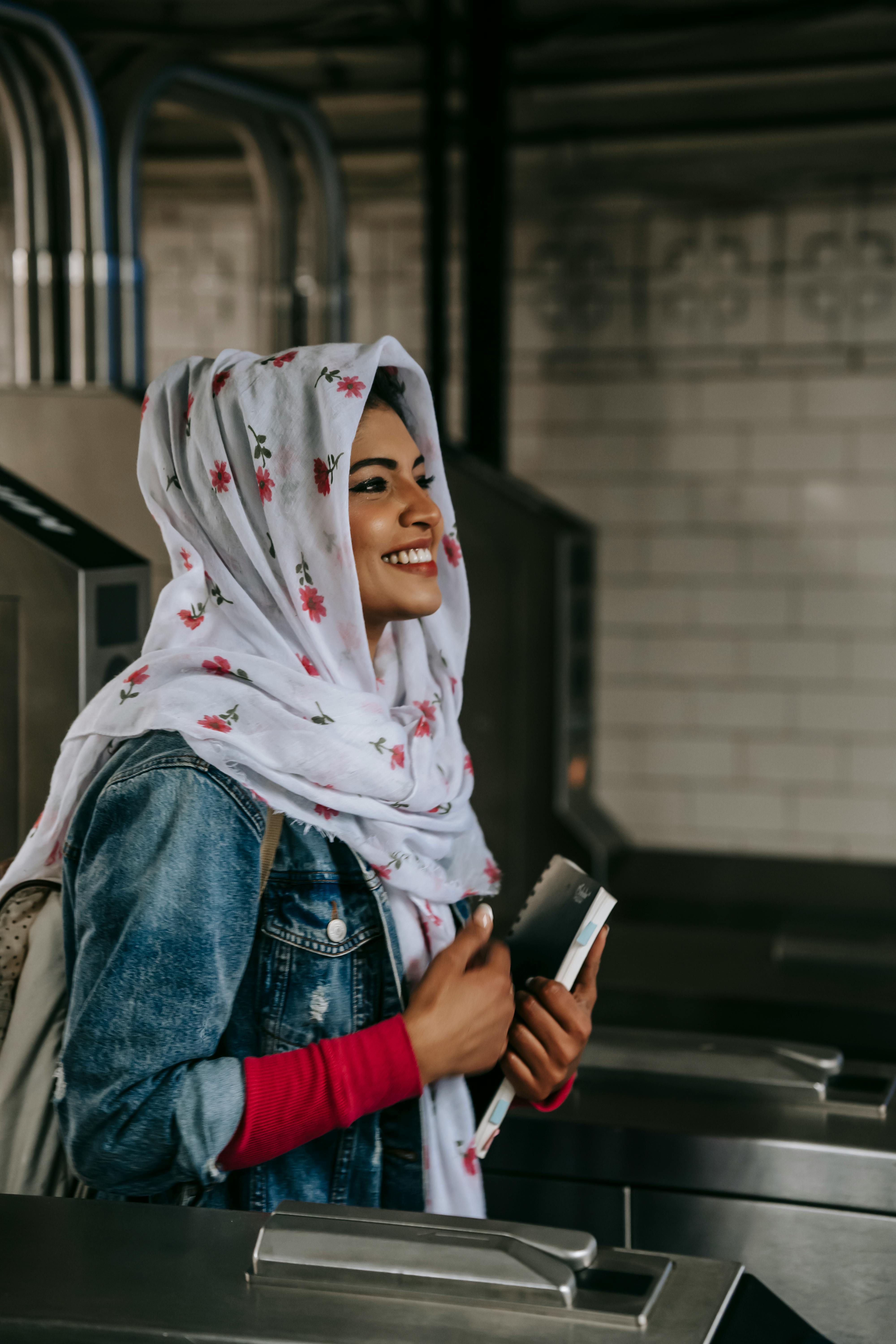 Side view of young Muslim woman with copybook wearing jeans coat and floral hijab smiling while entering subway station through automatic turnstile