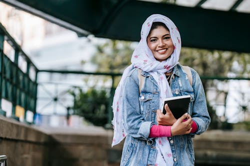 Free Content ethnic female in headscarf and denim coat with notebook looking at camera with toothy smile Stock Photo
