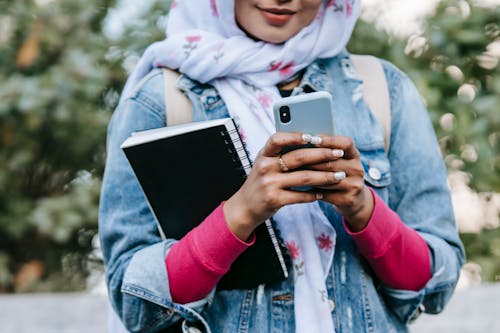 Crop anonymous female in headscarf and jeans jacket with rolled up sleeves and with notebook messaging on cellphone