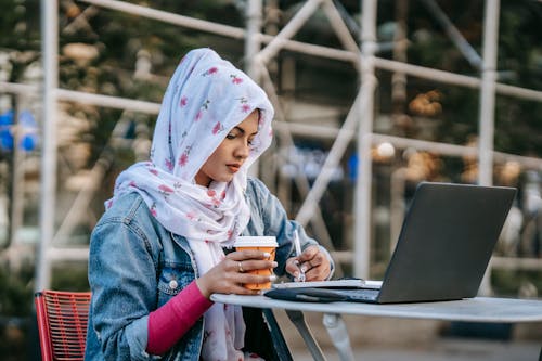 Focused Muslim female in headscarf and denim jacket taking notes from laptop while working and drinking coffee