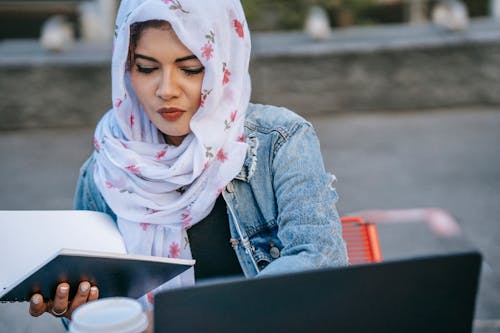 Gentle young ethnic female in headscarf working with notepad and netbook at table on blurred background of street