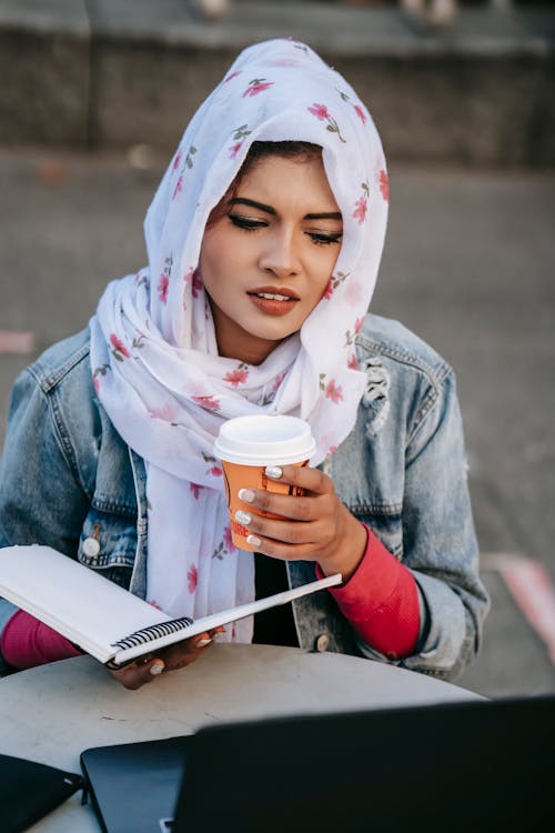 Young ethnic woman in headscarf with coffee studying