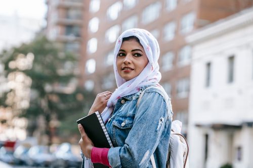 Free Calm Muslim female with dark hair and bright makeup in trendy clothes and headscarf standing with notebook on street in residential district in city in daytime Stock Photo
