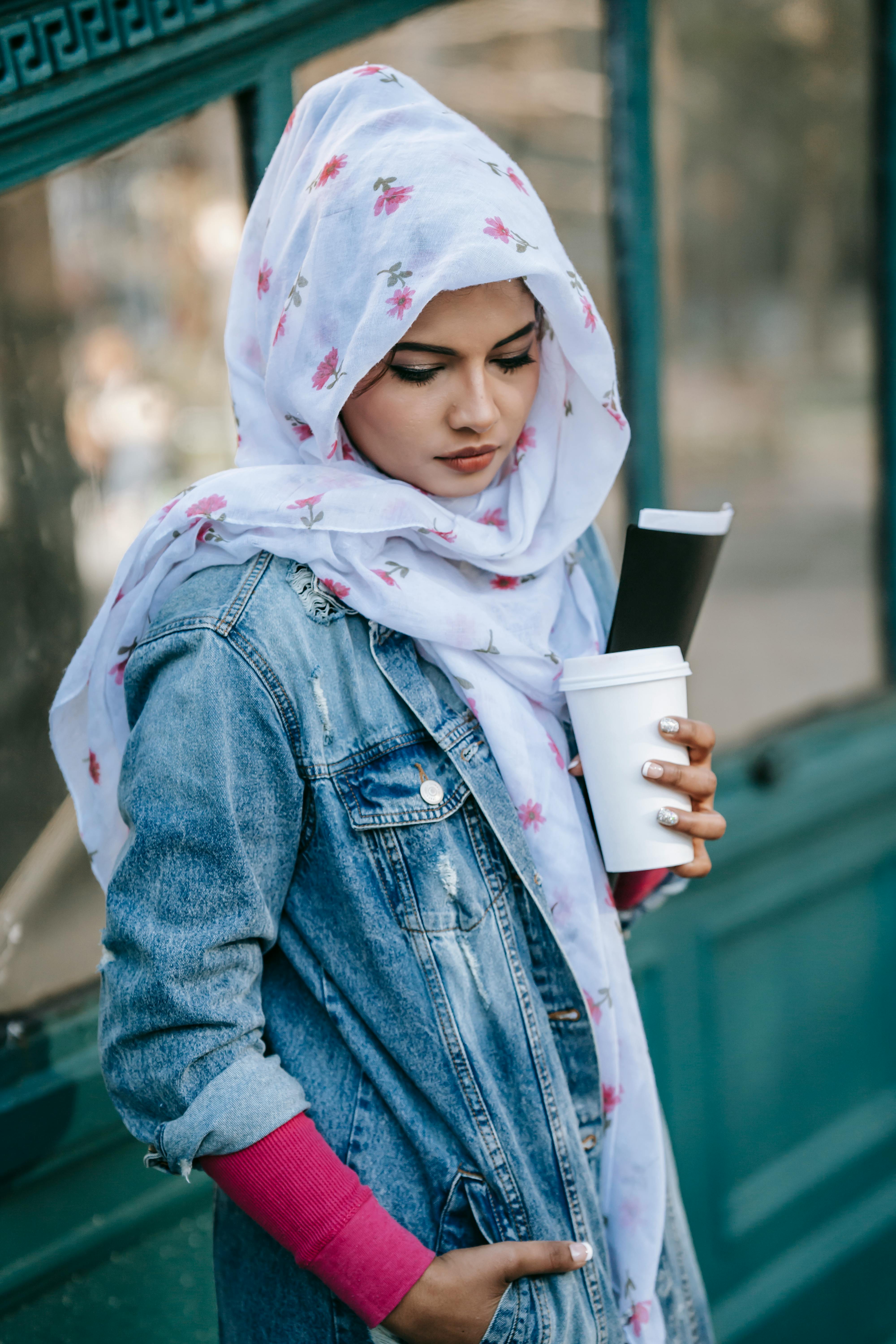 Tranquil Muslim woman in casual clothes and headscarf standing with cup of takeaway drink and folder with documents on street near construction with ornamental elements in daytime