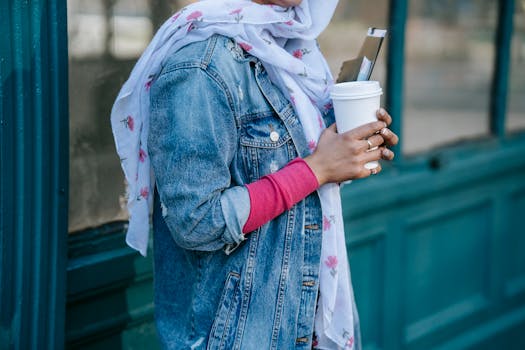 Crop anonymous woman in trendy outfit and headscarf standing with cup of takeaway beverage and notebook against construction with glass windows in daylight