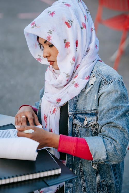 Side view of concentrated Muslim woman entrepreneur in casual clothes and  headscarf sitting at table and reading report on street in daylight