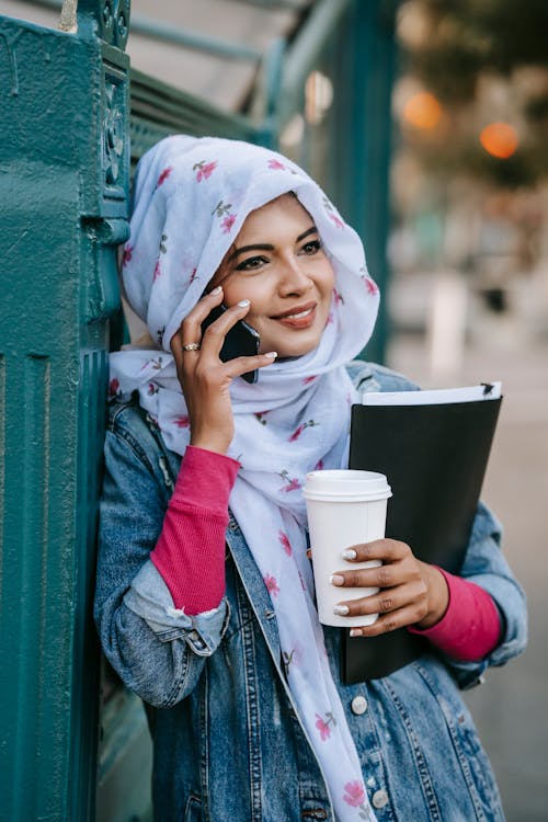 Free Cheerful Muslim woman in stylish clothes and headscarf standing with cup of takeaway coffee and folder with documents on street and speaking on smartphone in daytime Stock Photo