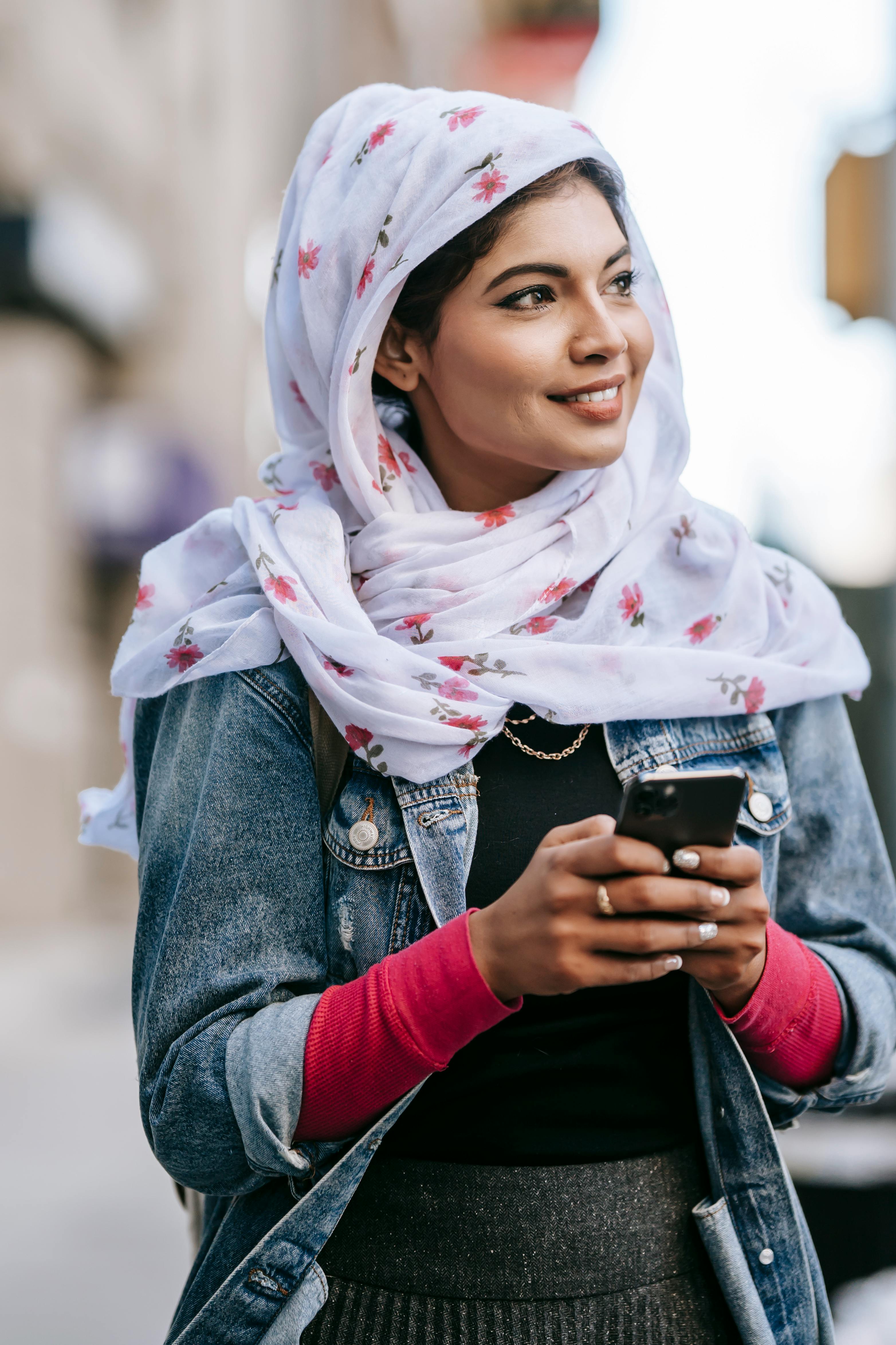 positive young lady wearing headscarf surfing smartphone while walking along street