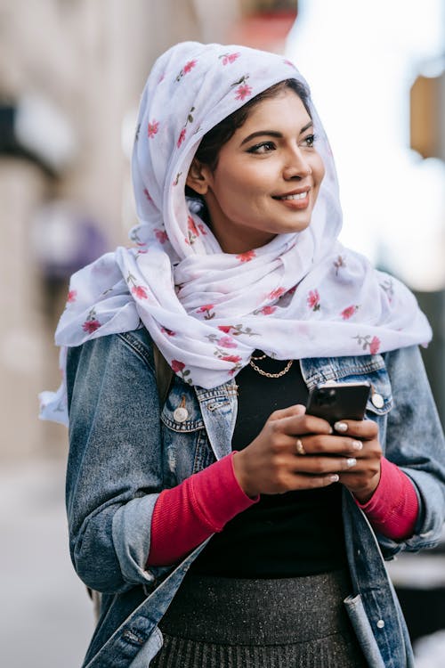 Positive young lady wearing headscarf surfing smartphone while walking along street