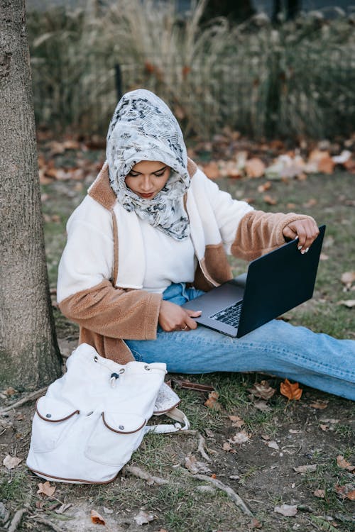 Concentrated young Muslim lady wearing floral hijab sitting on ground with backpack and remotely working on portable laptop located on knees in daylight