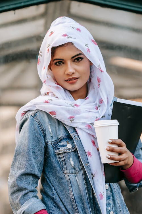 Ethnic woman wearing headscarf while standing with coffee to go and folder