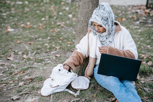 Concentrated young Muslim female freelancer in hijab leaning on tree in park while working on laptop and getting documents from white backpack