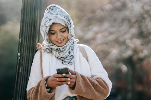 Cheerful young Muslim lady wearing warm coat and floral hijab leaning on pillar while surfing smartphone outdoors