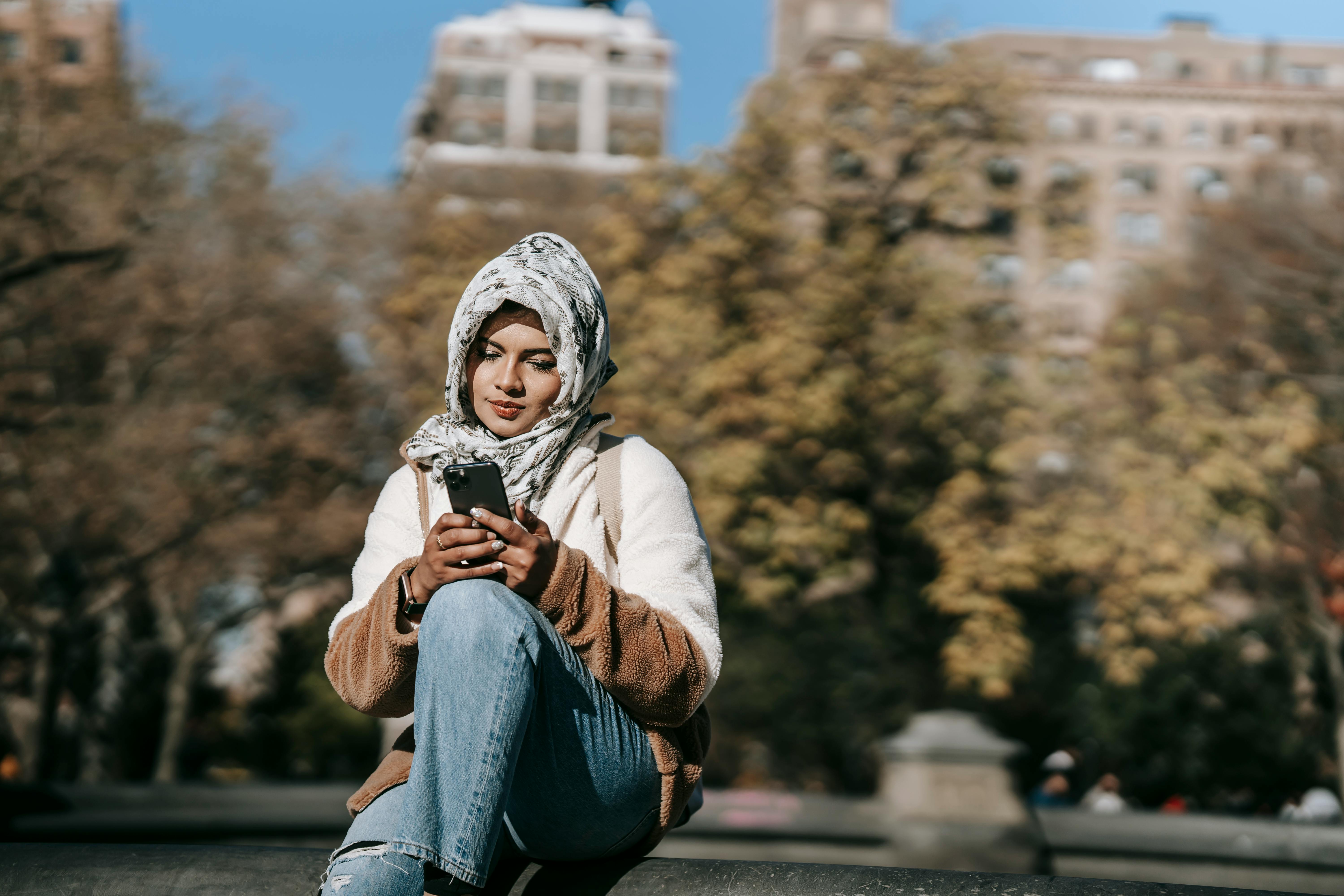 young ethnic woman using smartphone while sitting in city park