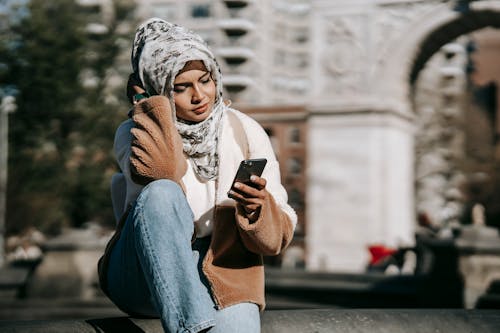 Free Stylish young Arab female in casual warm outfit and traditional hijab resting on city square and messaging on mobile phone on sunny day Stock Photo