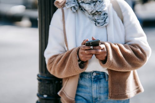 Crop unrecognizable woman in trendy warm outfit using mobile phone while standing on city street near metal pillar