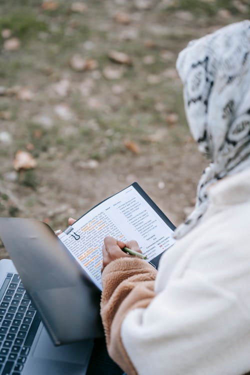 From above of crop unrecognizable Muslim female freelancer in casual clothes and headscarf reading and taking notes in documents while working remotely on laptop in autumn park