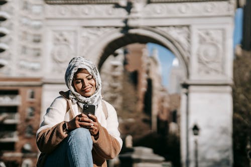Self assured young ethnic female in stylish clothes and Muslim headscarf smiling while messaging on smartphone sitting in city square near aged arch