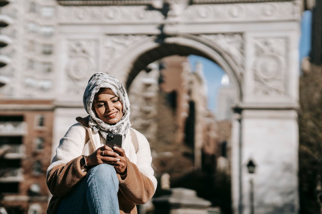 Free Self assured young ethnic female in stylish clothes and Muslim headscarf smiling while messaging on smartphone sitting in city square near aged arch Stock Photo