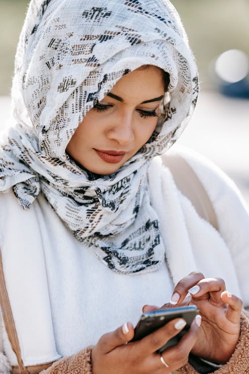 Crop young graceful Arab woman in casual outfit and elegant headscarf text messaging on smartphone on street in sunlight
