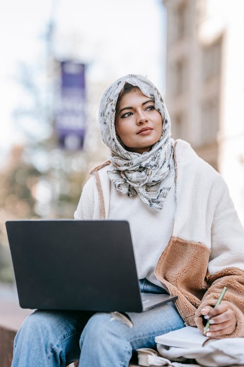 Young Muslim female freelancer in trendy clothes and hijab looking away thoughtfully while taking notes and using laptop during remote work on city street