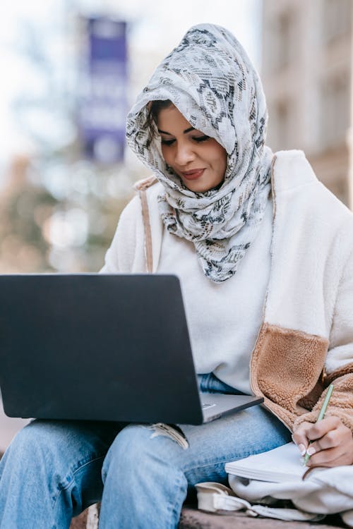 Positive young Arab female student in stylish clothes and headscarf smiling and taking notes in copybook during online lesson on laptop sitting on bench on city street
