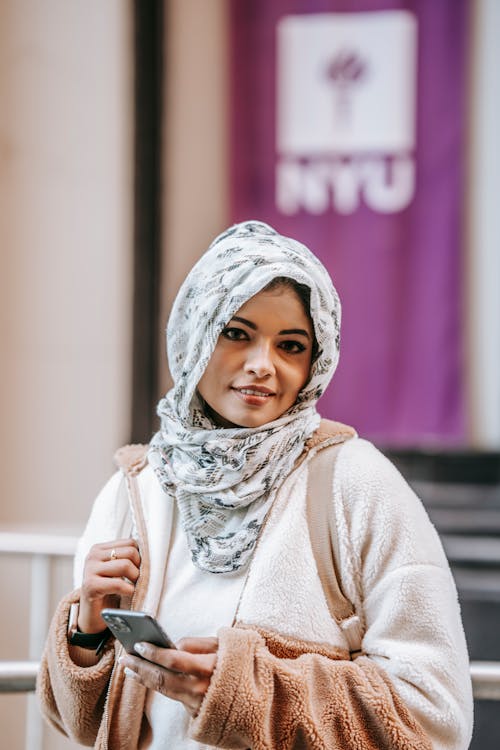 Self esteem young Muslim female millennial in trendy outfit and traditional headscarf smiling and looking at camera while browsing smartphone on city street