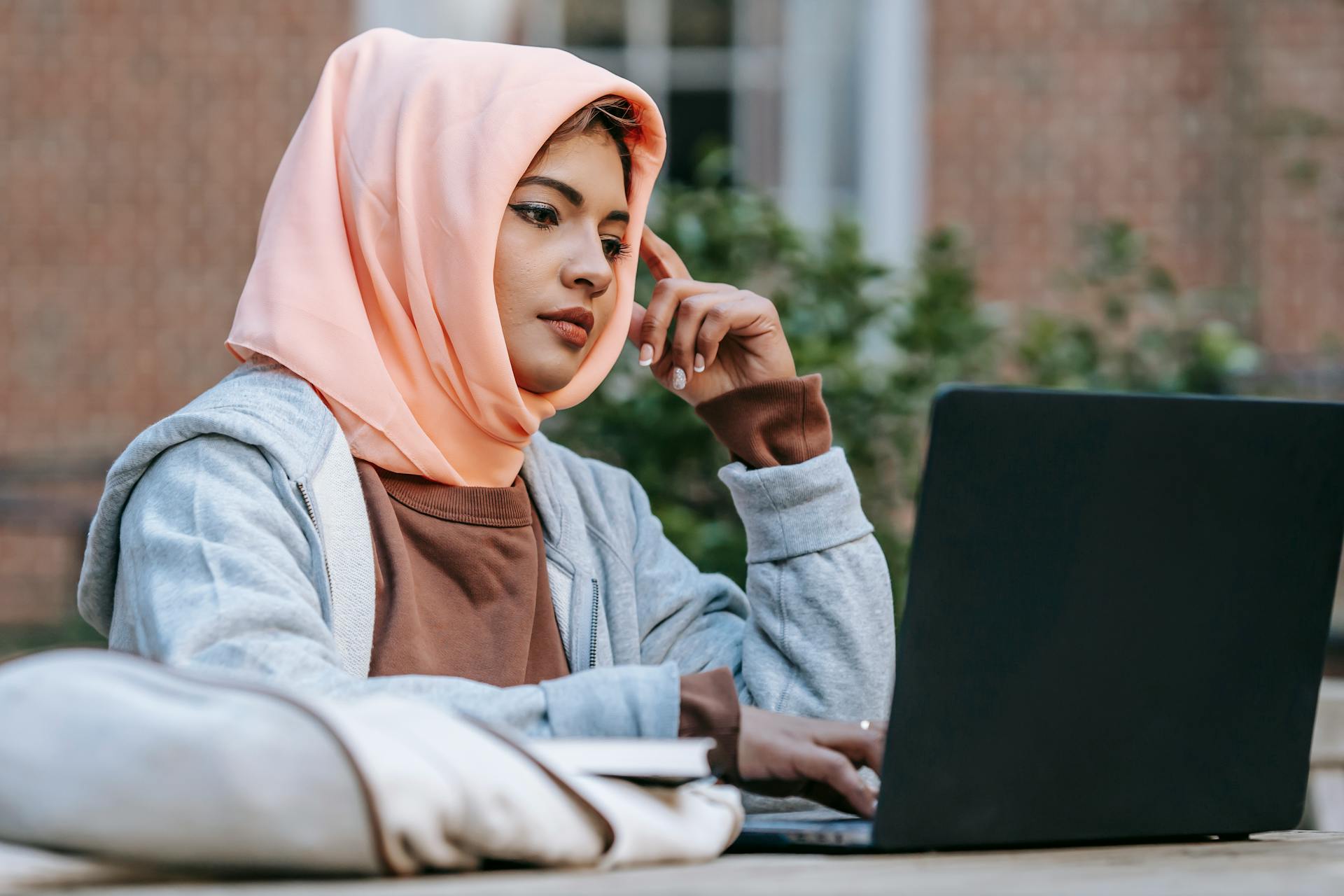 Pensive young Muslim female student touching head while using laptop during exam preparation in campus