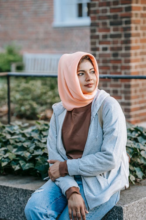 Free Content young stylish Muslim woman in casual clothes and hijab resting on city street and looking away dreamily Stock Photo