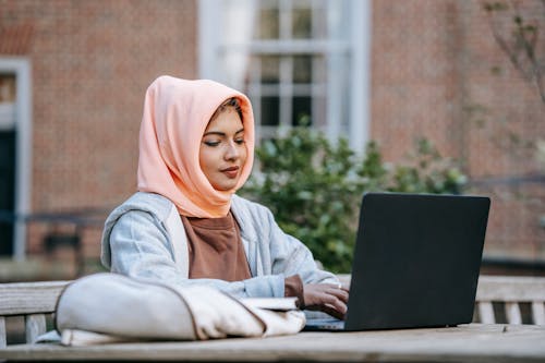 Stylish young Muslim female student typing on laptop at table in garden