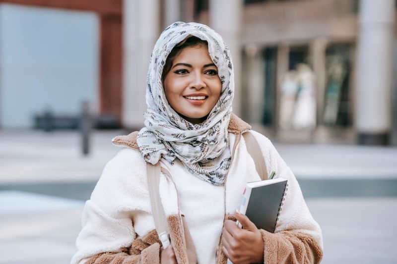 Happy young ethnic female student in casual clothes and hijab smiling and looking away while standing on street with notebook in hand after university